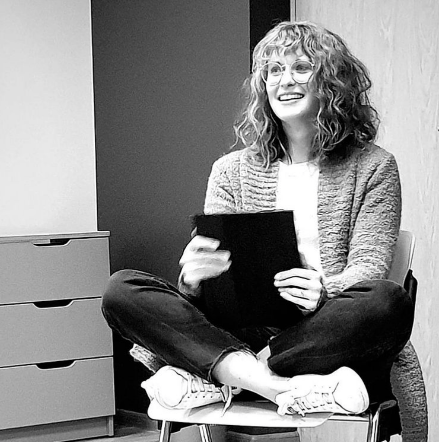 Callie leads a Bible Study at the Lighthouse. Here, she sits cross-legged in a chair holding an iPad and smiling.